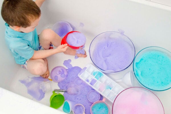 A child in a bathing suit sits in a bathtub. There are bowls of purple, blue, and pink bubble foam around him.