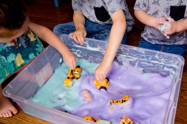 Three kids sit around a sensory bin filled with turquoise and purple bubble foam. They are pushing trucks through the foam.