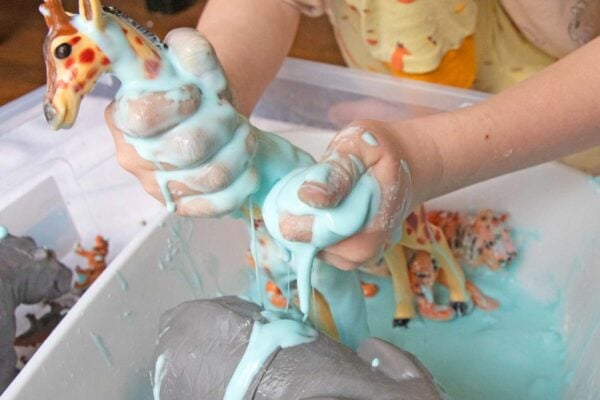 A child plays with blue oobleck in a sensory bin. There are also plastic animals in the oobleck.