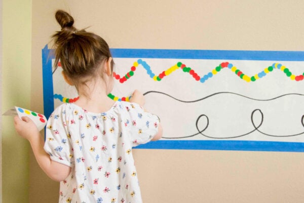 A child stands at a wall with a white piece of long paper on it. There are black lines. The child is placing stickers on the lines.