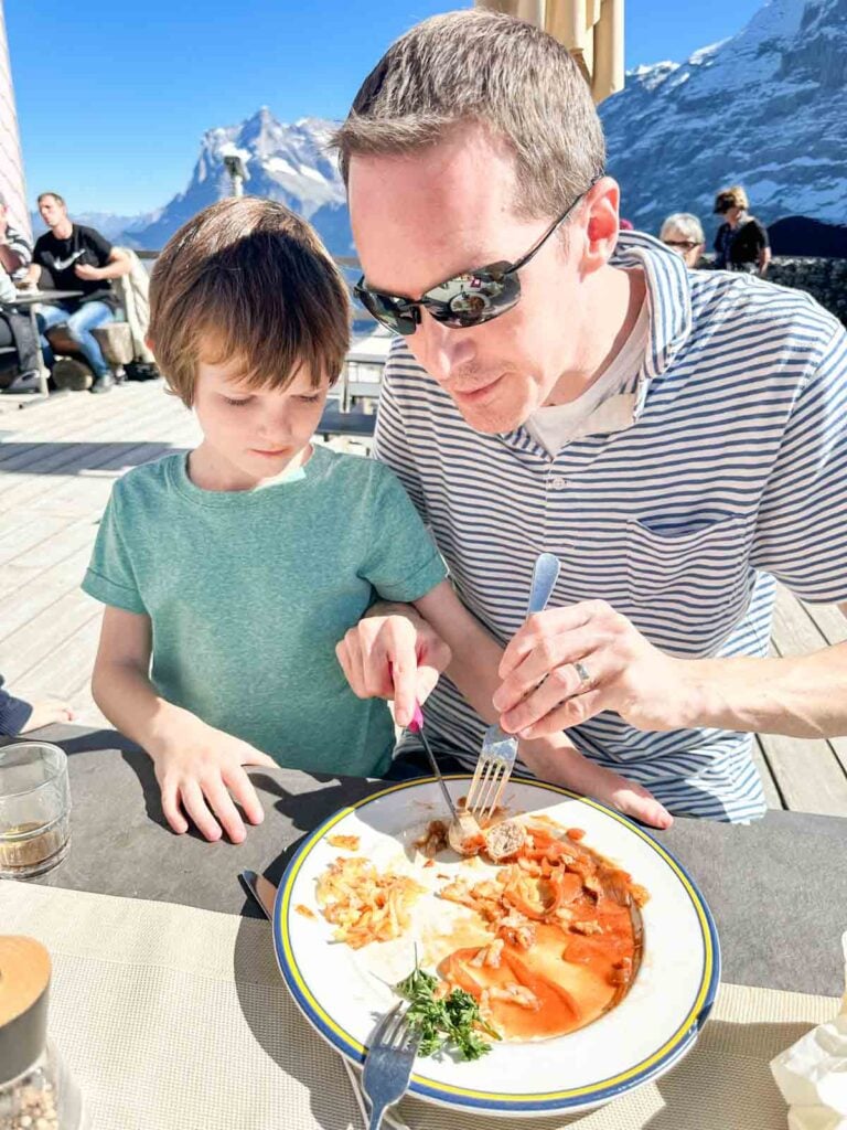 A parent shares their food with a child at an outdoor restaurant in the Alps.