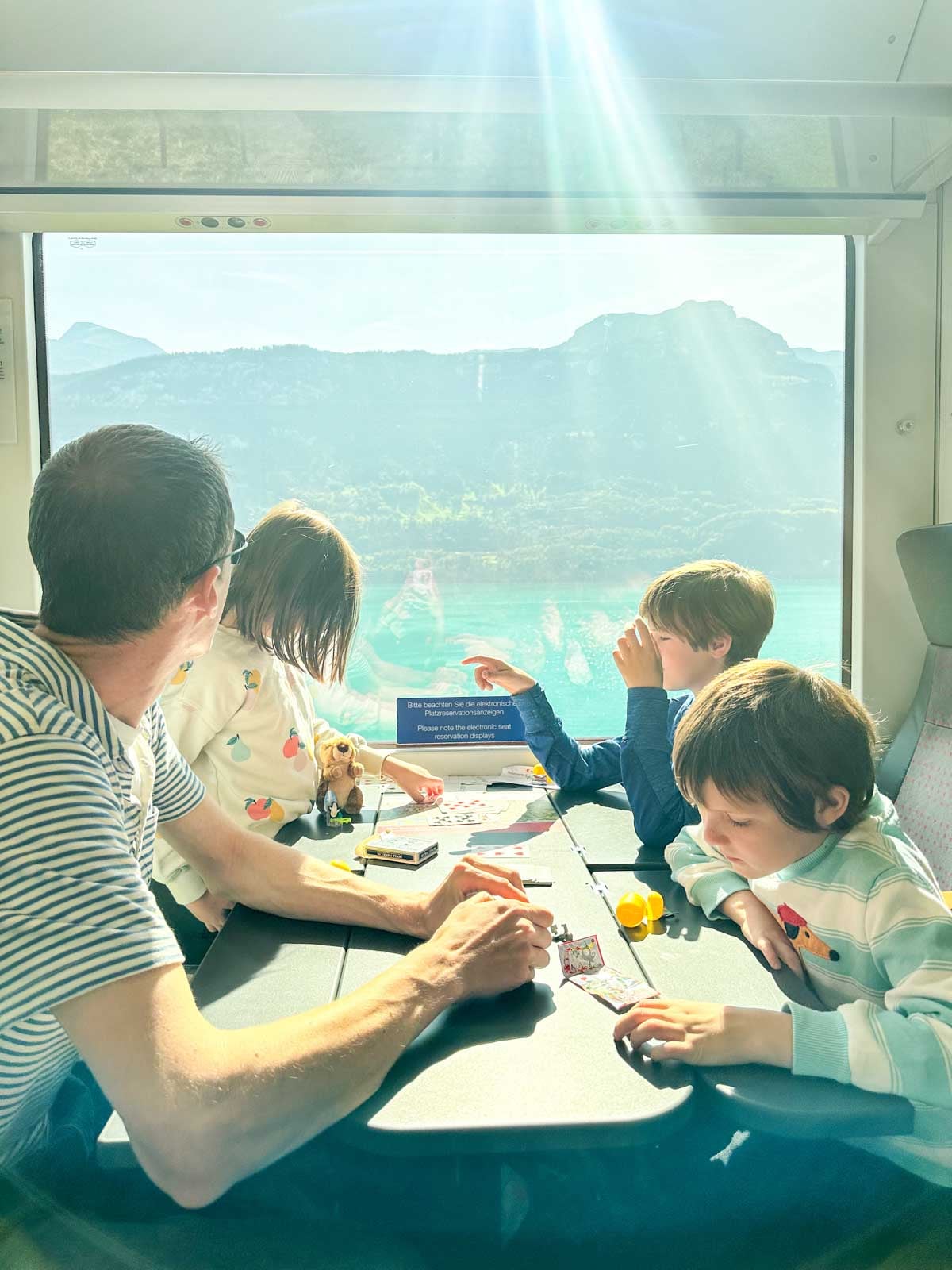 A man looks out a window onto a Swiss lake. His children are at a table with him.
