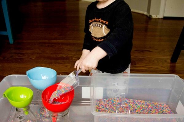 A child scoops rainbow rice into a red funnel. He is 2 years old and wearing pajamas.