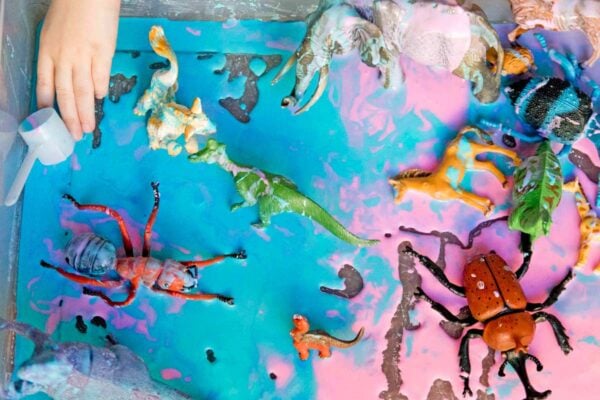 A sensory bin of pink and turquoise oobleck is in frame with several plastic animals. Two kid hands are working with the oobleck.