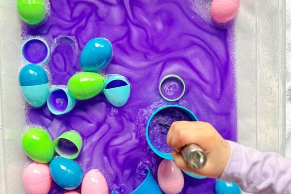 A child with a whisk is stirring purple metallic water in a sensory bin with Easter eggs.