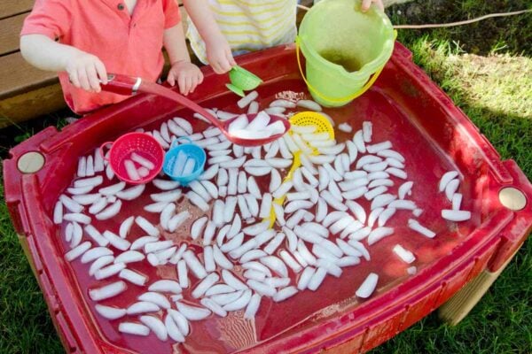 Two children play at a sand table full of ice cubes and water. One is scooping ice while the other holds a bucket.