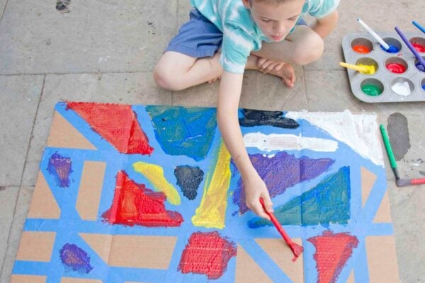 A child leans over a tape resist art project using red paint to fill in a triangle made of blue tape.