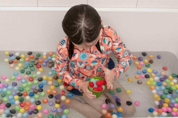 A child sits in a white bath, playing with red and green pom poms in a cup. The bath is filled with multicolored pom poms, making it a vibrant sensory activity for kids.
