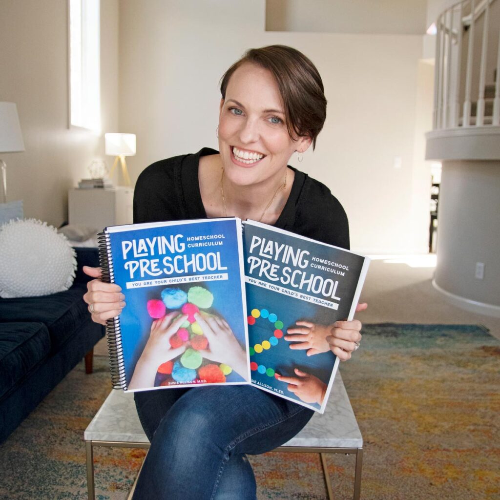 Author Susie Allison holds copies of her Playing Preschool homeschool preschool program. She is sitting in a living room.