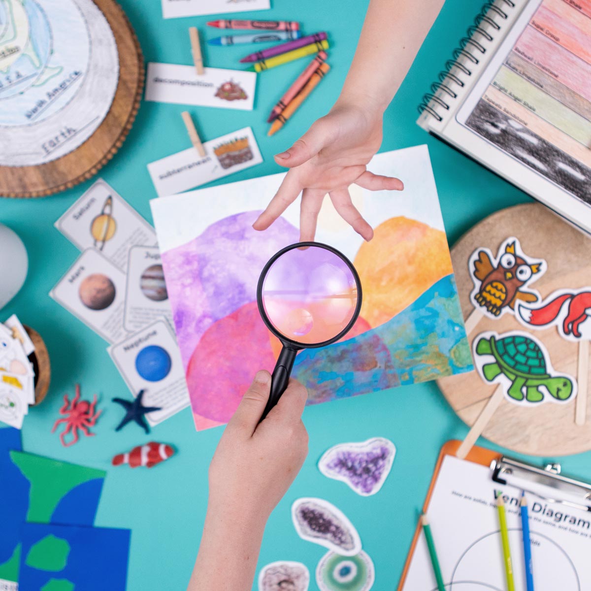 A child's hand reaches for a magnifying glass being held by another child. In the background, homeschool materials like crayons and projects are blurry.