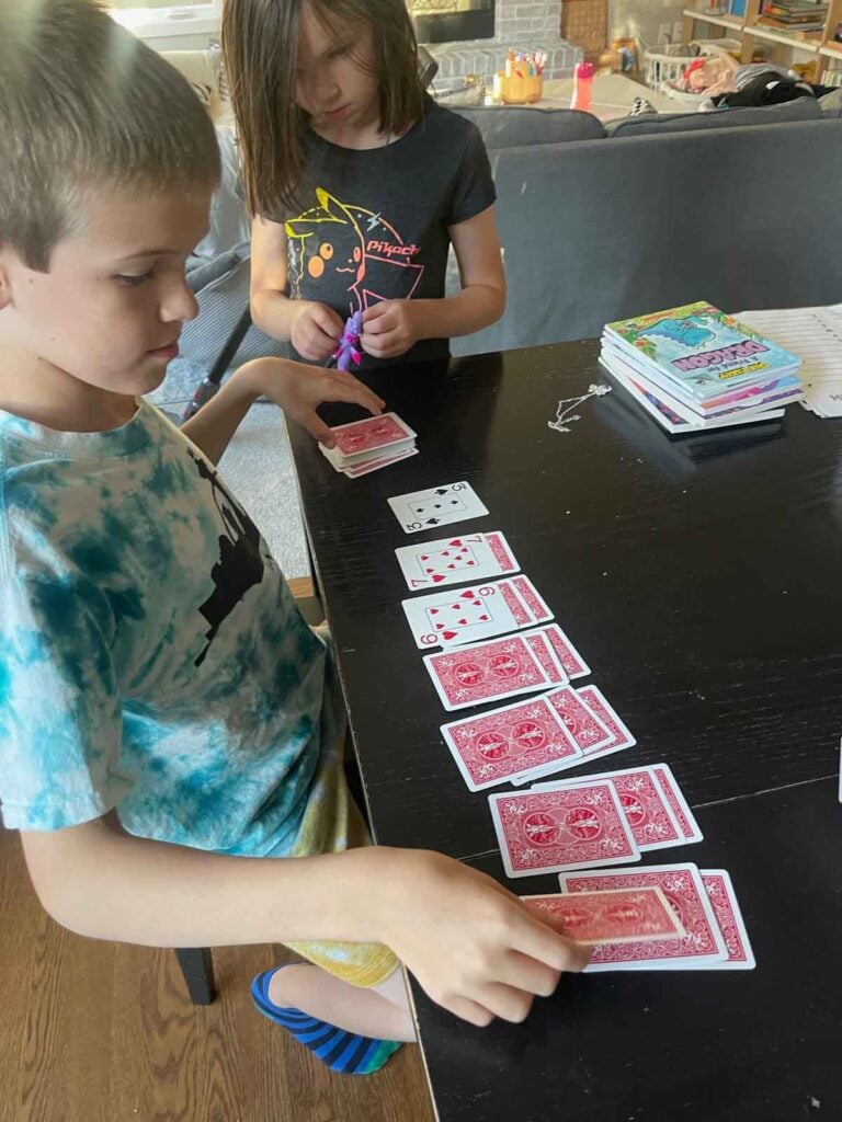 A child learns solitaire with his sibling. Cards are on a black kitchen table.