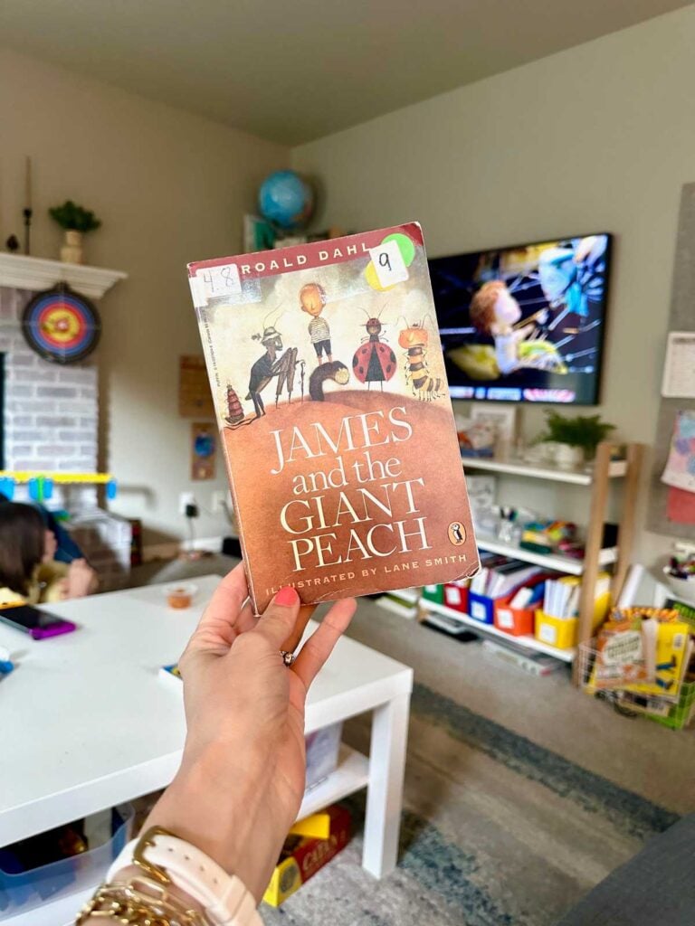 A woman holds a copy of James and the Giant Peach in front of a TV which is playing the movie version.
