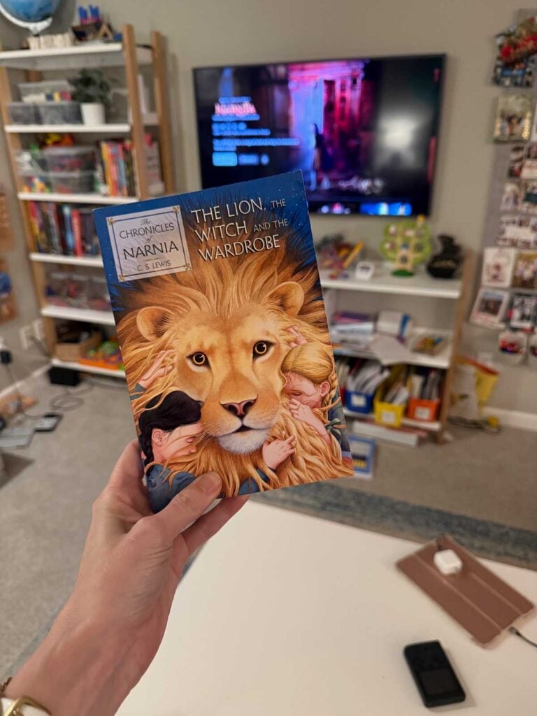 A woman holds a copy of the Lion, the witch, and the wardrobe in front of a TV playing the movie version.