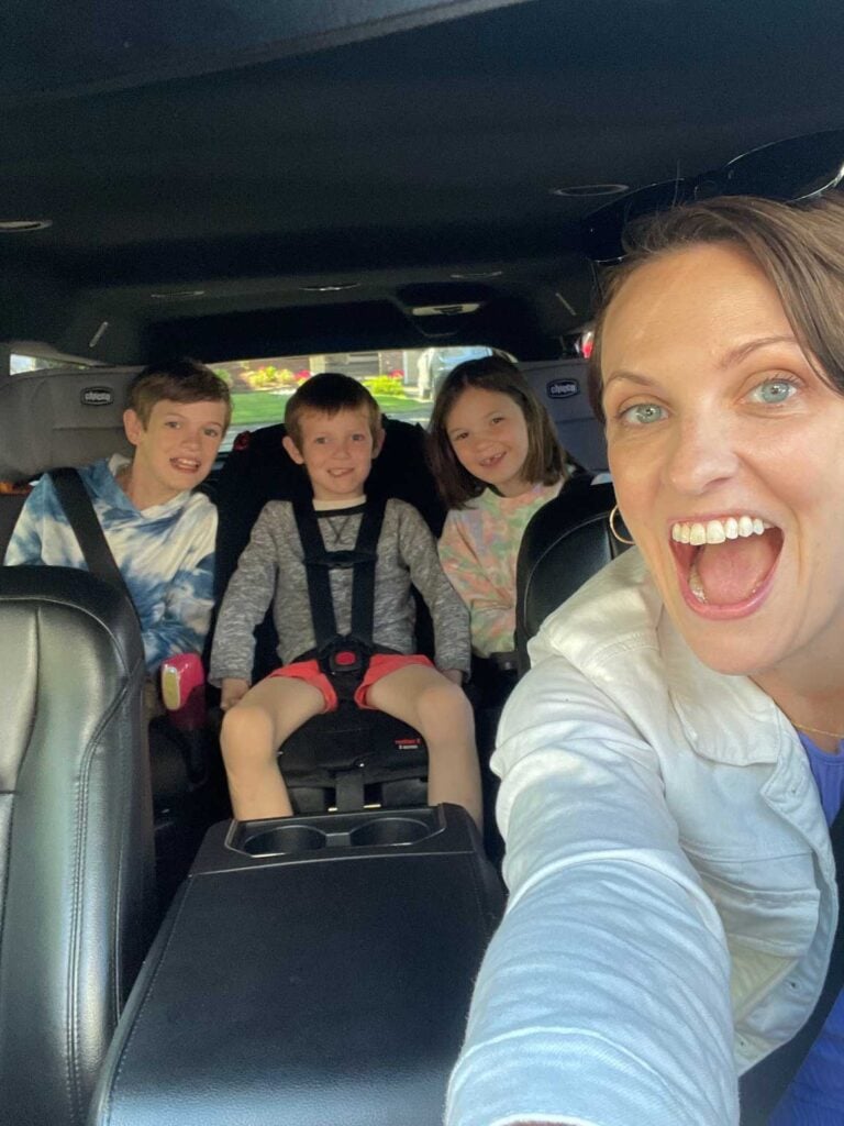 A mom in the front seat drives three kids in car seats. THey are all smiling.
