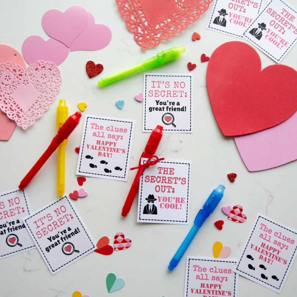A flat lay on a white counter has spy themed Valentines and invisible ink pens. The pens are rainbow colored with hearts on the counter.