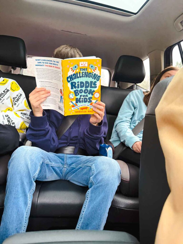 A tween reads a riddle book in the backseat of a car.